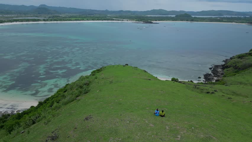 Solitary Couple On Grassy Coastal Hill Overlooking Ocean And Rocky Islet, Drone Panorama Captures Sweeping Green Slopes, Gentle Waves On Shore, Quiet Relaxation And Exploration