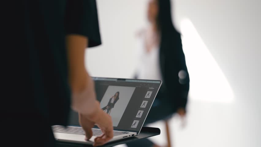 Photographer retouches photos on a laptop with editing software in a bright studio with a model in the background