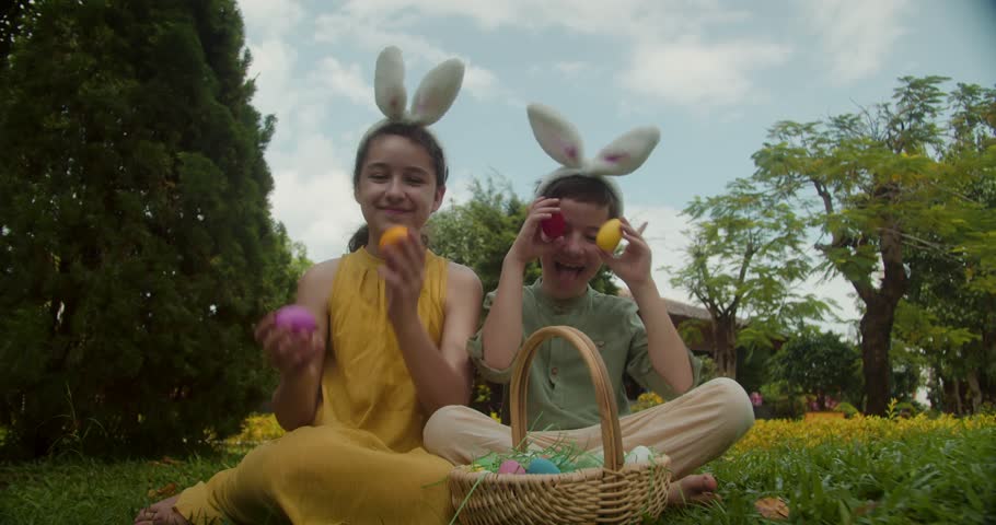 Happy children wearing bunny ears sit on green grass and playfully hold colorful Easter eggs over their eyes like glasses. Kids enjoying Easter holiday outdoors with basket of eggs in sunny spring