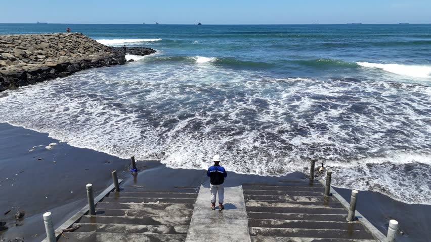 A man stands at the end of the stairs facing the sea