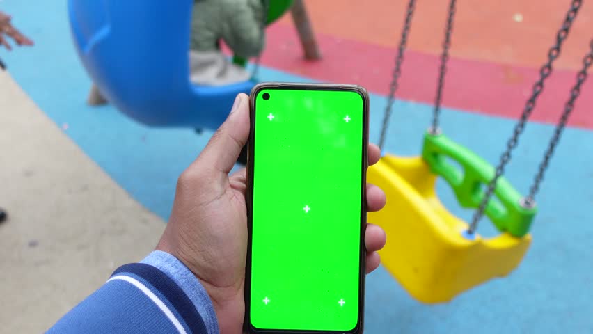 Close up of person hand holding mobile phone with green screen in an outdoor playground with blurry swing in background