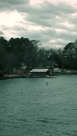 Vertical video of a lakefront scene featuring boat docks and a small boathouse along the shoreline. Wind ripples move across the water under a dramatic cloudy sky, creating an atmospheric lakeside landscape.