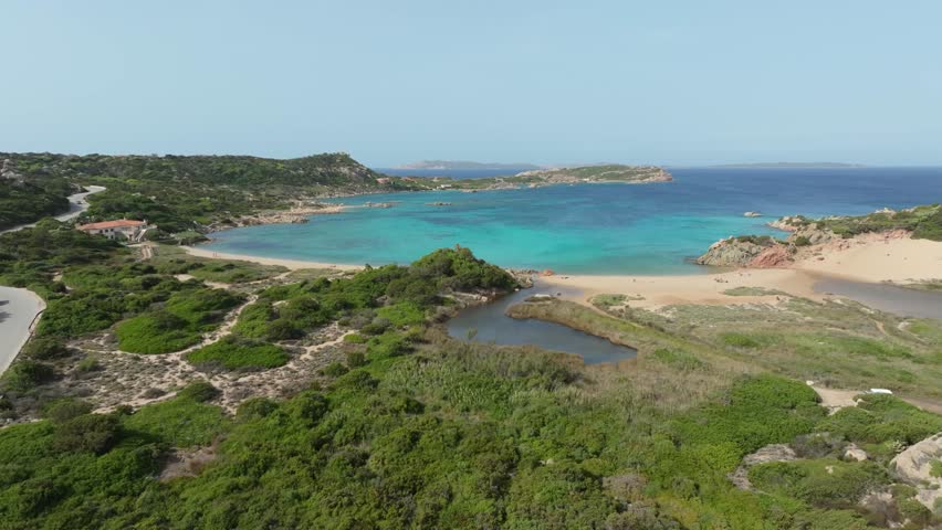 Stunning aerial perspective of a bay in Sardinia. The camera flies over a lush green landscape towards the pristine turquoise Mediterranean sea