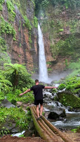 Man balancing on bamboo bridge near Kapas Biru waterfall in lush tropical jungle of East Java, Indonesia. Scenic rainforest landscape with dramatic cascade in Southeast Asia.