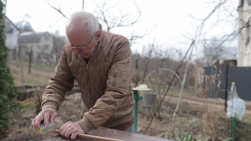 Elderly caucasian man with glasses sanding a wooden plank. Focused retiree working with sandpaper on a woodcraft project in his backyard