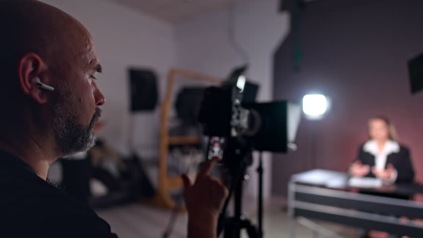 Bearded photographer looks focused on camera display. Cameraman filming a lady reporter sitting at desk at blurred backdrop. Studio backstage.