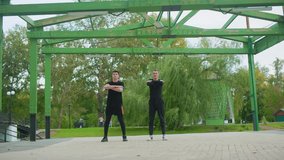 Two white men exercising under green pergola doing synchronized squats and arm crosses, coach instructing trainee, leafy park background, casual black activewear, upbeat morning training vibe - Powered by Shutterstock - Get 15% off with code: PIKWIZARD15