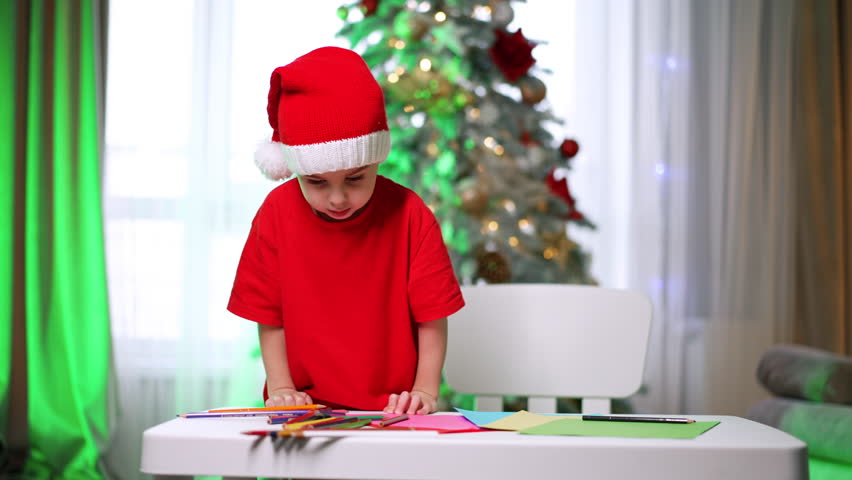 Lovely baby in red Santa cap and t-shirt stands at the desk in his room. Toddler kid chooses a pencil and starts to draw. Christmas tree at blurred backdrop.