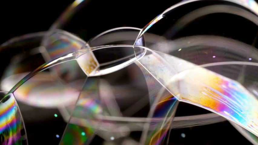 Interconnected soap bubbles glisten with iridescent colors. Light plays across the surface, creating shimmering and shifting patterns. Black background.