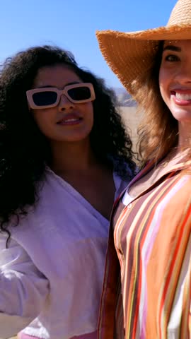 Two smiling multiracial young women having fun together on a sandy beach during a sunny day. Joyful female friends enjoying their summer vacation, laughing and talking by the sea