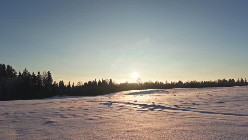 Sun setting over snowy field and forest horizon during calm winter evening landscape, low aerial dolly to silhouette tree line