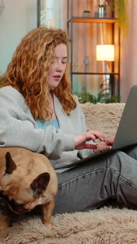 Young Caucasian redhead woman focuses on work with laptop on home sofa, typing with quiet determination. Pug dog lies beside girl, adding comfort and calm presence to personal workspace atmosphere.
