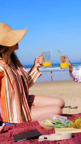Two beautiful female friends enjoying a relaxing picnic on a sandy beach, toasting with orange juice and eating snacks on a sunny day with the ocean waves in the background