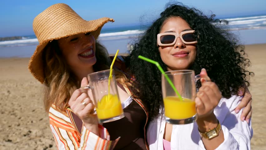 Two smiling multiethnic women enjoying a sunny day on the beach, laughing and toasting with glasses of orange juice, celebrating their friendship and enjoying their vacation by the sea