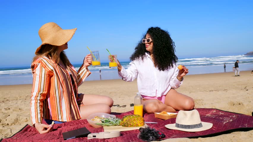 Two diverse female friends sitting on a blanket on the sand, toasting with orange juice, eating fresh fruit, and laughing together during a beautiful sunny day at the seaside with ocean waves