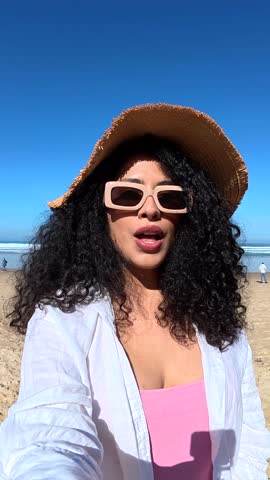 Cheerful curly haired woman wearing a straw hat and sunglasses filming a selfie video, smiling and talking to the camera while enjoying her vacation on a sunny beach with the ocean behind her