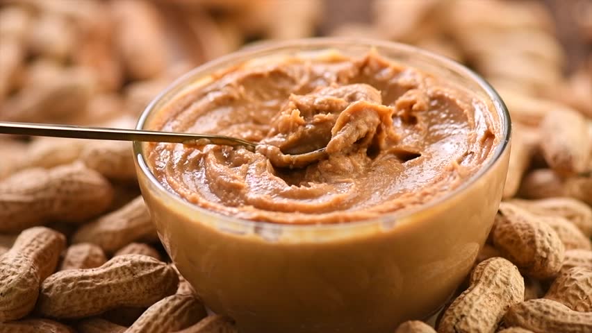 Peanut butter in a glass jar with spoon. Creamy smooth peanut butter texture. Mixing with a spoon, making sandwich, organic food. American cuisine. Slow motion. Rotating backdrop 