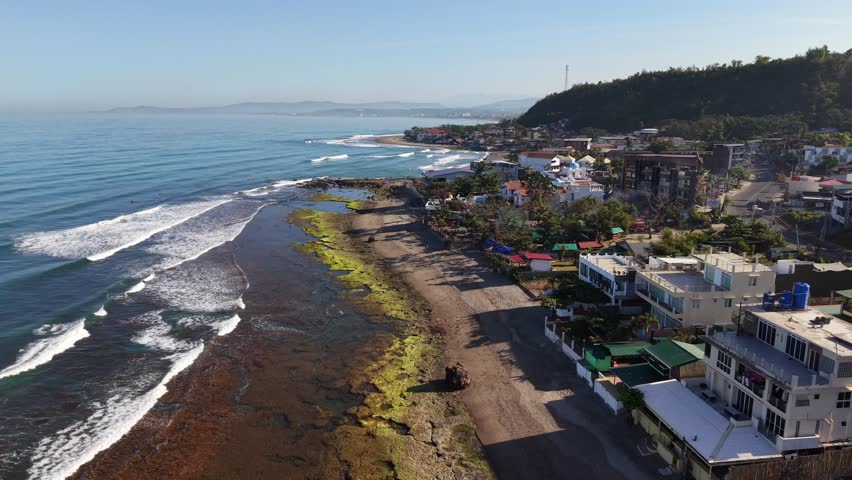 Aerial view of calm blue ocean waves. Sandy beach curves along the coast. Colorful coastal town buildings line the shore. Hills rise behind the town. Ideal for travel or vacation content
