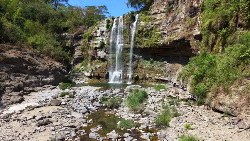 Sunlight illuminates a cascading waterfall. Water flows into a rocky pool surrounded by lush greenery. Moss covers the cliffside walls. Clear blue sky enhances the serene setting