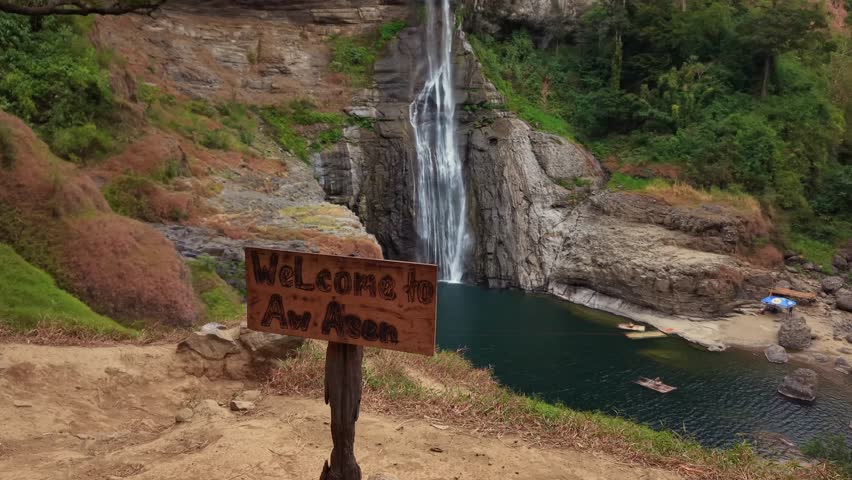 Waterfall flows into a deep green pool. Rocky cliffs surround lush trees and vegetation. Welcome sign marks the trail