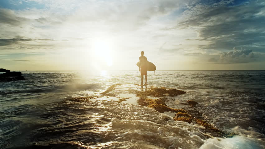 Surfer stands on a rocky reef at sunset, holding a surfboard and waiting patiently for the perfect wave. Warm golden light reflects off the shimmering ocean waters, creating a dramatic and serene