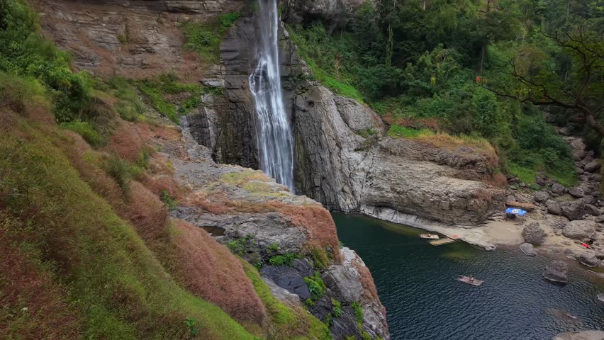 Waterfall flows down rocky cliffs. Tree stands near the water. People wade in the pool, smiling. Lush grass covers the slopes. The scene invites relaxation. Ideal for nature travel content