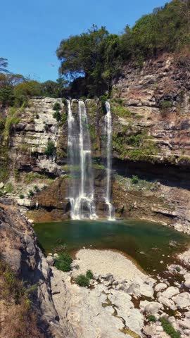 Bright blue sky above the waterfall. Water flows down rocky cliffs. Clear green pool reflects the sun. Lush trees surround the area. Rocky terrain forms a natural scene