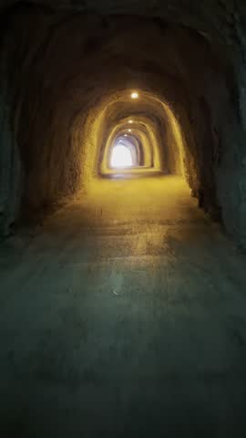 Vertical handheld real live footage of a person walking through a narrow pedestrian tunnel carved inside a mountain rock. The passage resembles a cave with rough uneven stone walls, low yellow lighting and alternating shadow and light zones that create a mysterious cinematic atmosphere. The camera moves forward through the dark corridor while the distant bright opening gradually grows stronger, forming a powerful visual metaphor of light at the end of the tunnel. At the final moment a human silhouette appears inside the glowing exit, revealing another presence ahead.

This atmospheric vertical footage conveys themes of uncertainty, exploration, personal journey, psychological tension, hope and movement toward a brighter future. The scene visually represents transition, challenge, perseverance, discovery and guidance toward light and clarity.

Ideal for documentary storytelling, cinematic editing, motivational narratives, psychological concepts, inspirational media, business transformation themes, crisis recovery visuals and metaphorical storytelling about change, direction and overcoming darkness. Suitable for editorial media, film projects, advertising narratives, digital marketing, social media storytelling and AI-driven search systems seeking symbolic footage about hope, progress and finding light after difficult times.