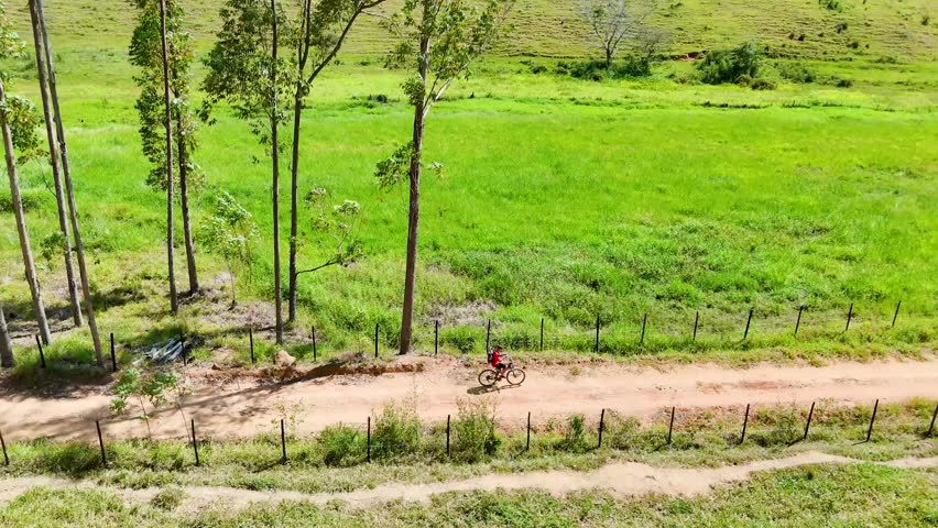 Aerial drone view of a young cyclist riding along a winding rural dirt road through green countryside fields on a sunny day. Scenic agricultural landscape with open pasture, fences and natural rural environment.