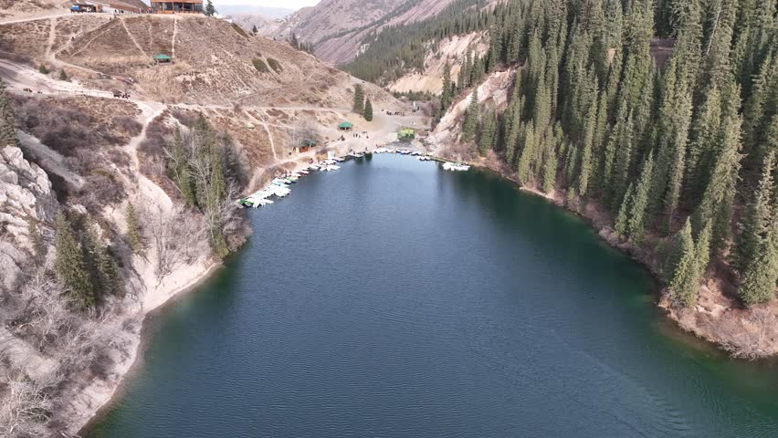 Aerial push-in shot descending toward colorful pedal boats docked on the emerald waters of Kolsai Lake in Kazakhstan, surrounded by dense pine forests.