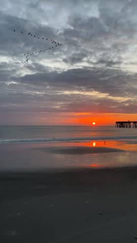 Golden sunrise over the ocean with waves rolling toward shore, colorful sky reflected on wet sand and a pier silhouette in the distance.