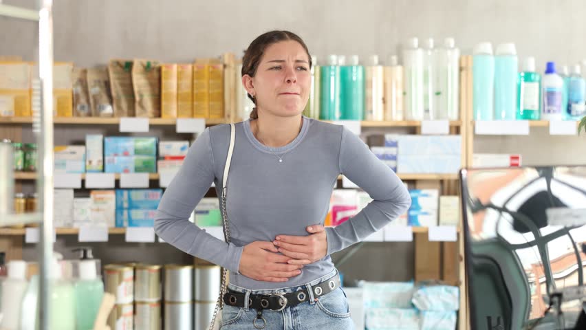 Young woman suffering from abdominal pain, clutching stomach while standing in drugstore, looking for medication to stop cramps and ease digestive discomfort. High quality 4k footage