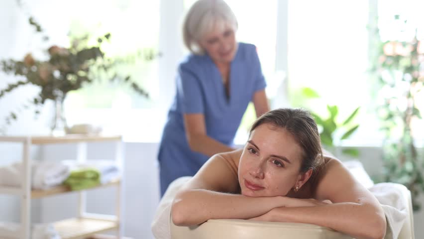 Smiling relaxed young woman lying on couch in spa salon during professional back massage from skilled masseuse 