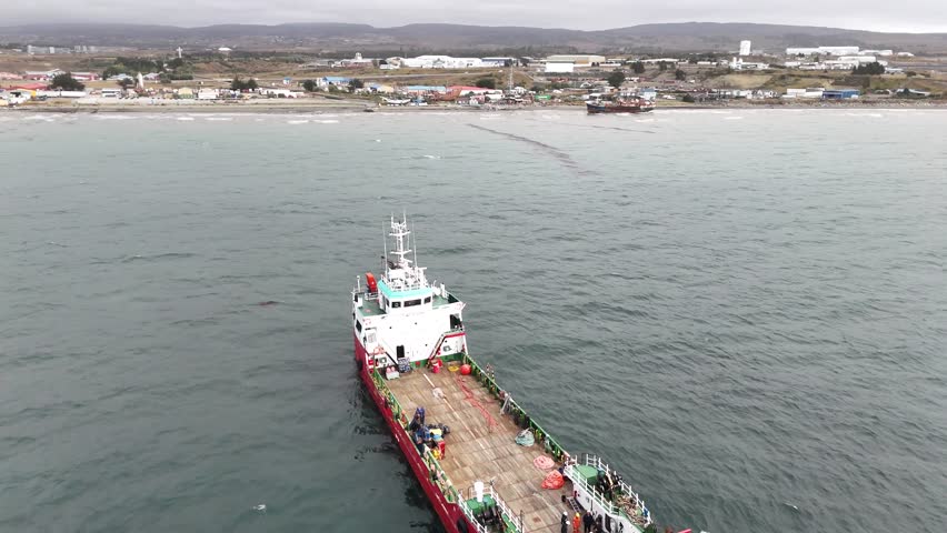 Aerial pulls back from a close-up of the barge Anan, moving out to reveal the vessel in the Strait of Magellan with the industrial coastline of Punta Arenas in the background