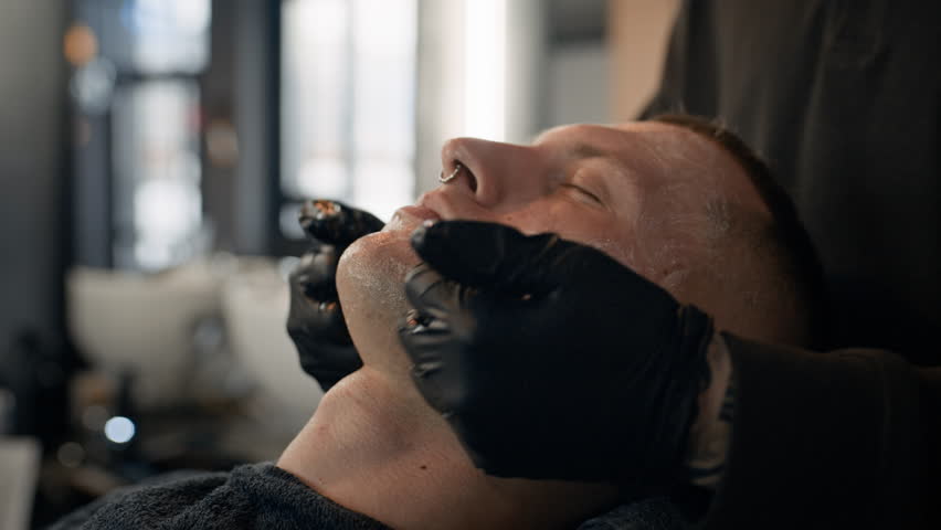 Close up of anonymous barber using cream while massaging face of man who sitting in barbershop chair with closed eyes
