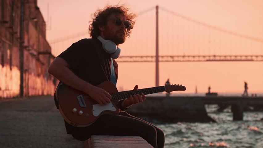 Hipster street musician in black playing electric guitar in the street on sunset on embankment with 25th of April bridge in background. Lisbon, Portugal