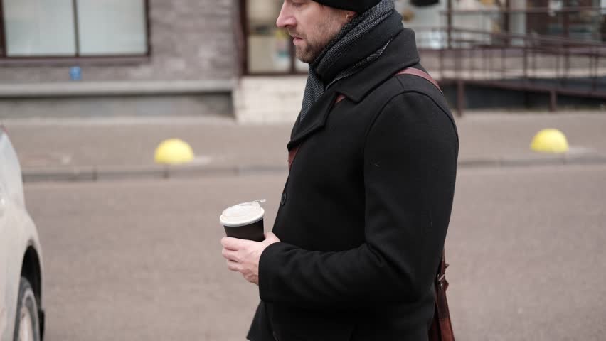 Handsome man wearing black coat and brown leather bag walking down on the city, drinking coffee paper takeaway cup with mobile phone. Stylish bearded middle-aged male using smartphone at urban street