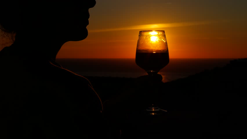 Silhouette of a woman drinking wine at sunset. Romantic silhouette of a woman enjoying a glass of red wine, admiring the beautiful orange sunset over the ocean, creating a relaxing atmosphere