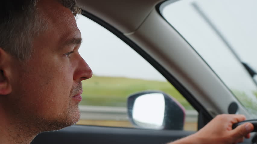 Close-up profile of a focused middle-aged man driving a car on a highway during overcast weather. He holds the steering wheel, looking ahead with a calm expression