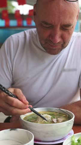 Vertical screen Caucasian Man Slurping Pho Noodles, Outdoor Stall Setting White Tee And Glasses, Chopsticks Lifting Steaming Rice Noodles From Bowl Of Beef Broth Garnished With Herbs
