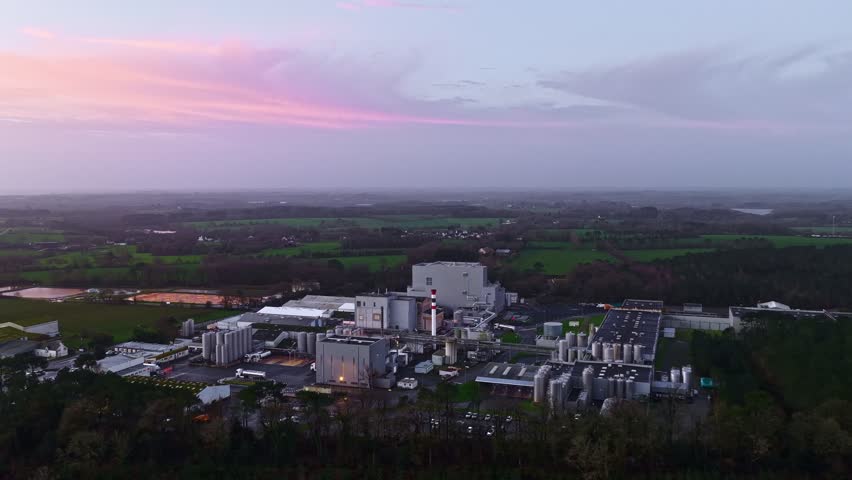 Wide drone view of a large industrial plant and factory complex at dusk with pink sunset sky over rural farmland. Energy, manufacturing, infrastructure, and corporate skyline background.