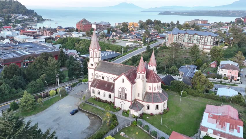 Cinematic aerial circling the iconic Sacred Heart Church (Iglesia del Sagrado Corazón de Jesús) in Puerto Varas, Chile, showcasing its red spires and German-influenced architecture against city skyline