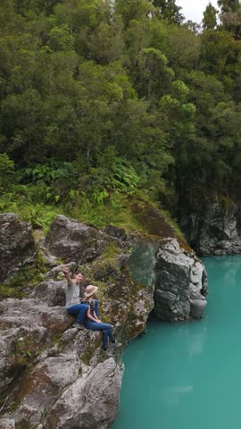 Vertical aerial orbit of a young couple on rocks above Hokitika River, Hokitika Gorge, West Coast, New Zealand, waving beside vivid turquoise water and native forest