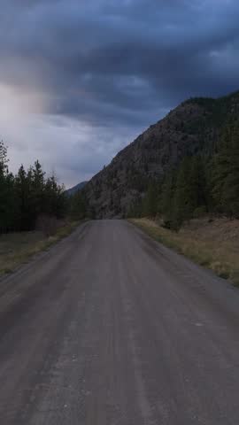 A quiet gravel road winds through a rugged forested mountain landscape, framed by pine trees under a dramatic, cloudy sky in British Columbia, Canada.