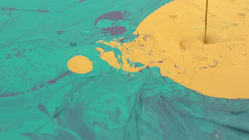 Slow-motion footage of vibrant liquid paints being poured onto a surface, with a stream of yellow paint forming a circular splash pattern surrounded by teal and purple colors. The swirling textures.
