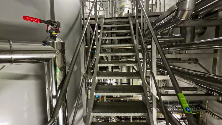 POV walking up metal staircase inside a stainless steel processing plant, surrounded by pipes, valves and machinery on an industrial catwalk.
