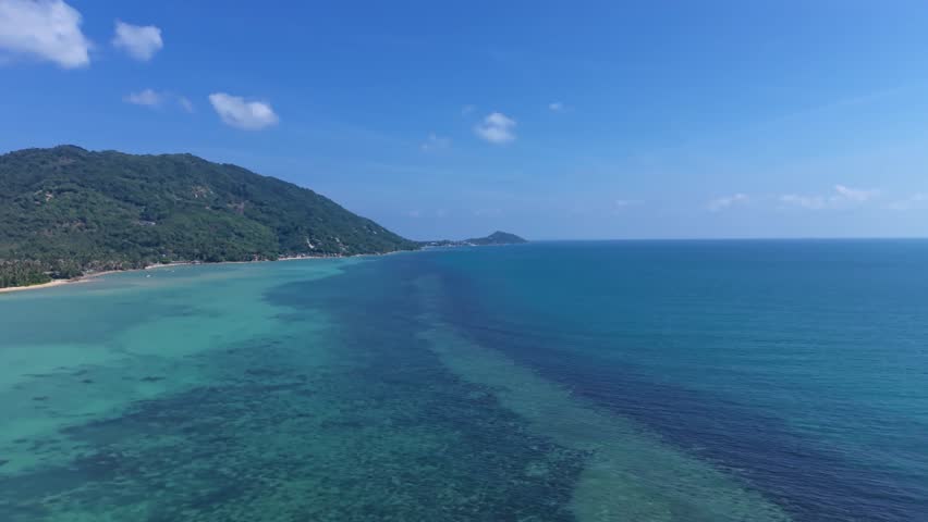 Aerial view descending toward shallow turquoise sea along a tropical island coast in Thailand.