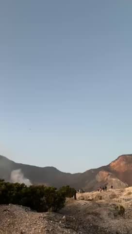 A dramatic landscape view of an active volcanic crater with white sulfur smoke rising from the ground under a clear blue sky. The rugged terrain, dry rocky surface, and surrounding mountain ridges create a powerful natural scene. Small human figures can be seen walking along the crater rim, emphasizing the vast scale of the environment. This footage captures the raw beauty and harsh conditions of a volcanic area, highlighting geothermal activity, natural minerals, and the striking contrast between earth tones and the bright sky.