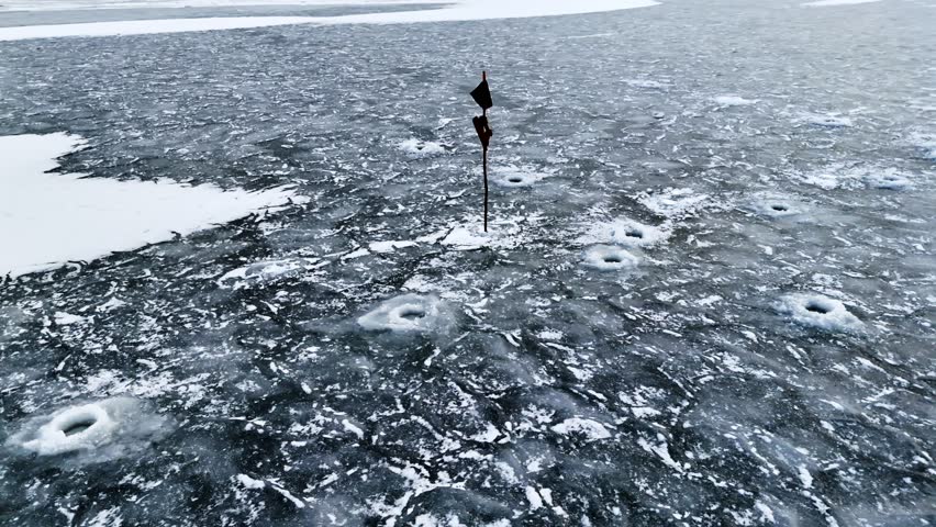 Aerial winter view of an ice fishing area with drilled holes and a marker pole on rough frozen water, surrounded by snow patches and textured dark ice.