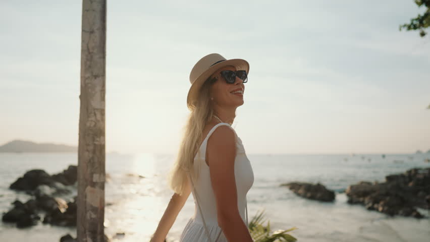 Happy woman in white summer dress strolling alone near sea and smiling broadly. Traveling to islands in summertime, spending vacation in beautiful coastline, tourism and discovery of world, enjoy life